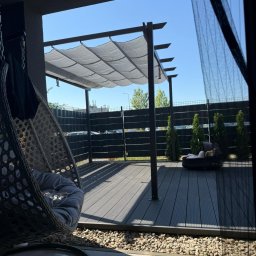 Gospodarstwo Ogrodnicze Krzysztof Dudek - Nowoczesny taras z zadaszeniem pergolą i wiszącym fotelem, wykończony deską kompozytową i kamieniami. W tle widoczny płot i zieleń.