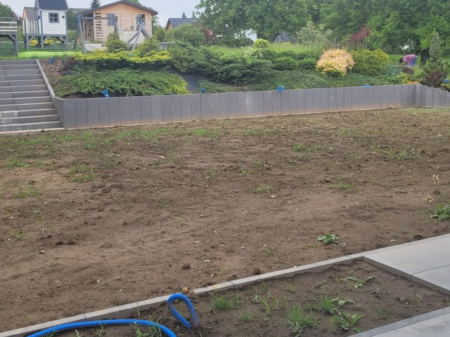 Przygotowany teren pod ogród z nową ścianą oporową i schodami. Widoczne elementy małej architektury ogrodowej w tle. Wąż nawadniający na pierwszym planie.