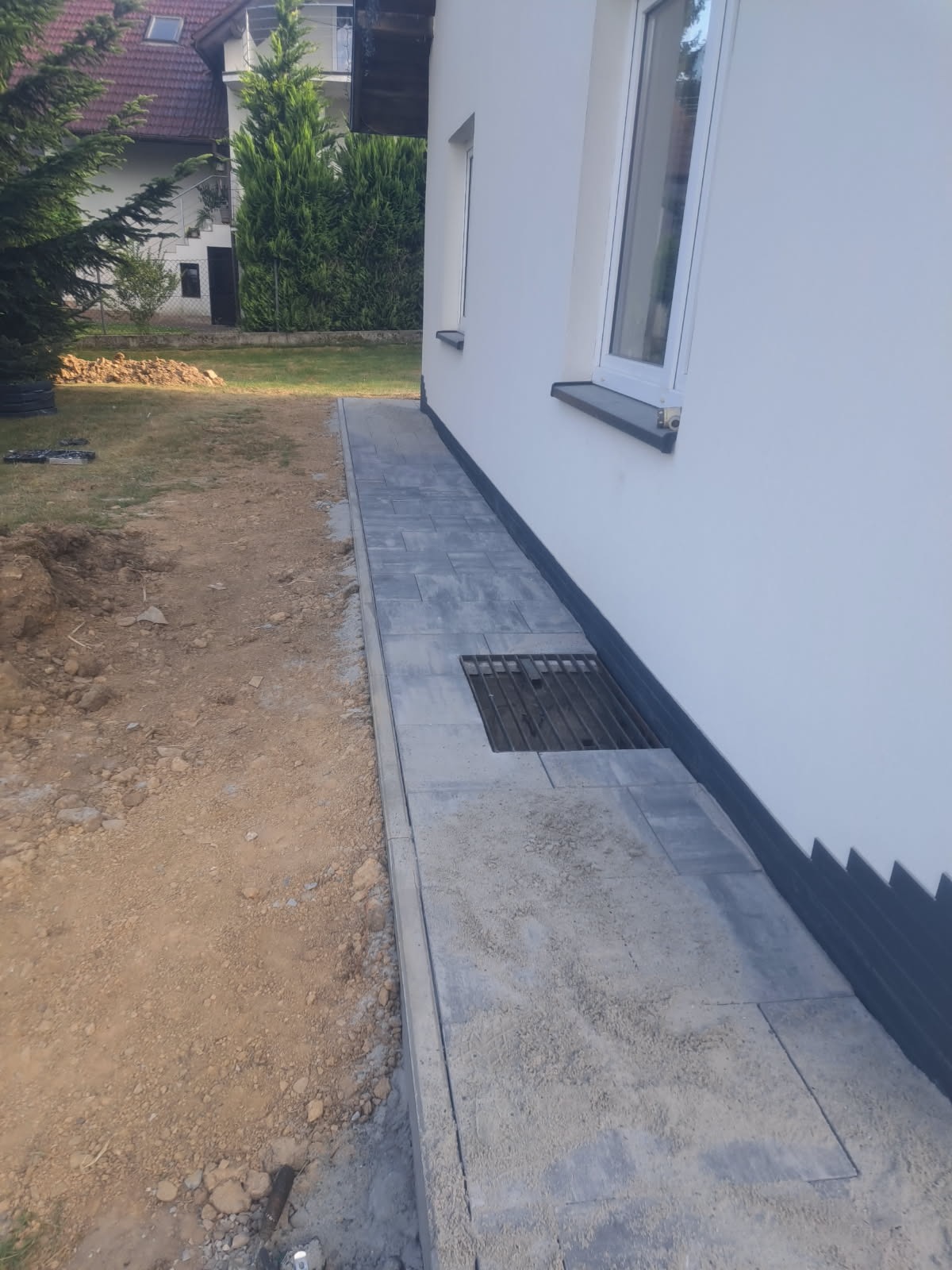 Ścieżka z szarej kostki brukowej ułożona wzdłuż białej ściany budynku, z kratką wentylacyjną wkomponowaną w nawierzchnię. Częściowo wykończona, obok ziemia.