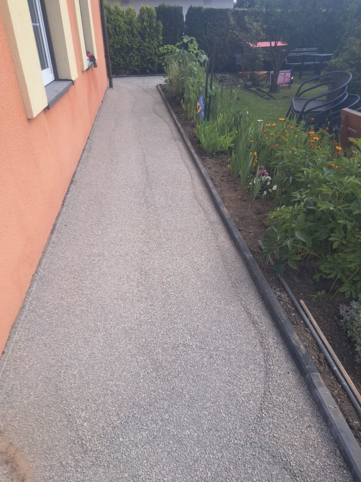 Ścieżka z szarego, drobnego kruszywa prowadząca wzdłuż budynku i rabaty kwiatowej. Widoczne narzędzia ogrodnicze i elementy wyposażenia ogrodu.