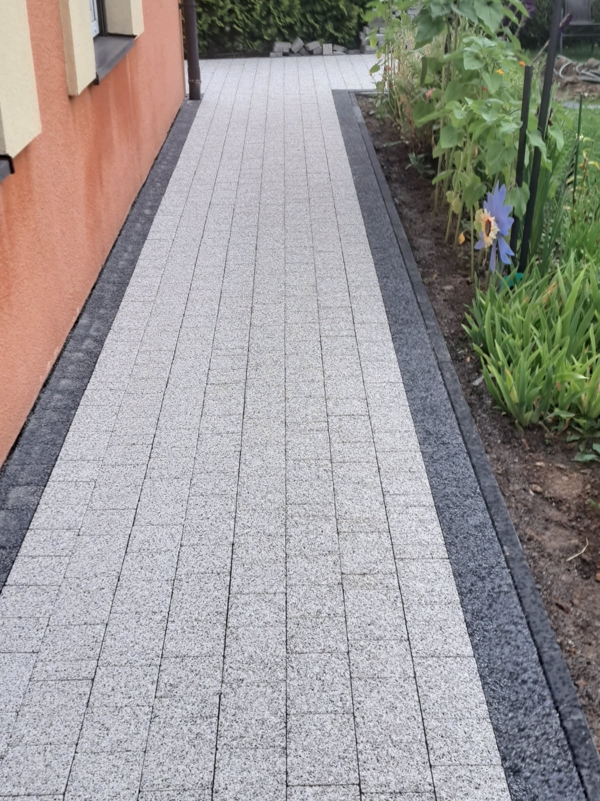 Jasnoszara kostka brukowa ułożona wzdłuż pomarańczowej ściany budynku, obramowana ciemniejszą kostką. W tle widoczna zieleń ogrodu i fragment rynny.