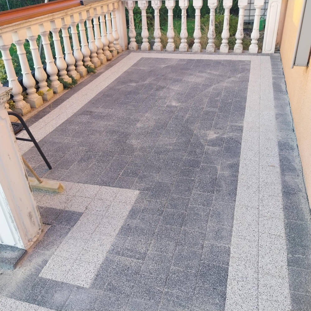 Szara kostka brukowa na tarasie z białym obramowaniem, balustrada z dekoracyjnymi elementami. Widoczny fragment krzesła i miotła. Układ wzoru prostopadły do balustrady.