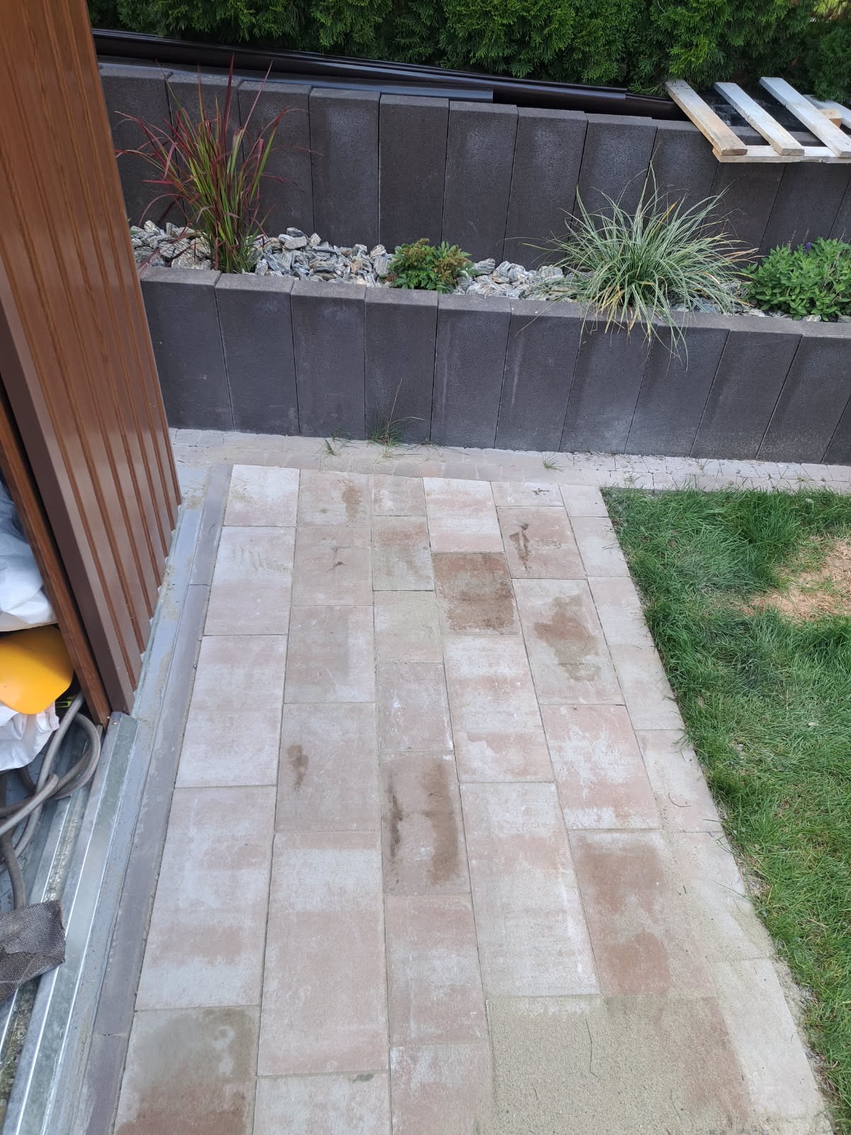 Ułożona kostka brukowa w ogrodzie, widok z góry. Szara kostka, trawa i rośliny w podwyższonej rabacie. Częściowo widoczne narzędzia i brązowe drzwi.