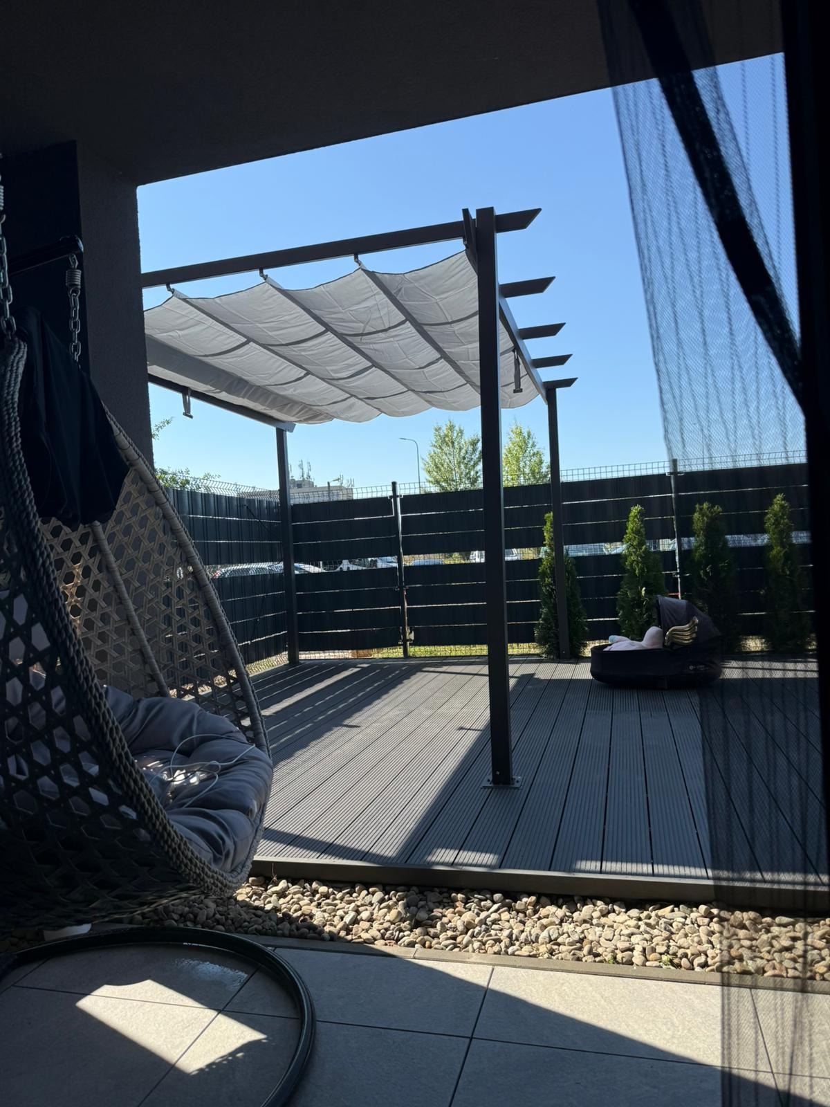 Nowoczesny taras z zadaszeniem pergolą i wiszącym fotelem, wykończony deską kompozytową i kamieniami. W tle widoczny płot i zieleń.