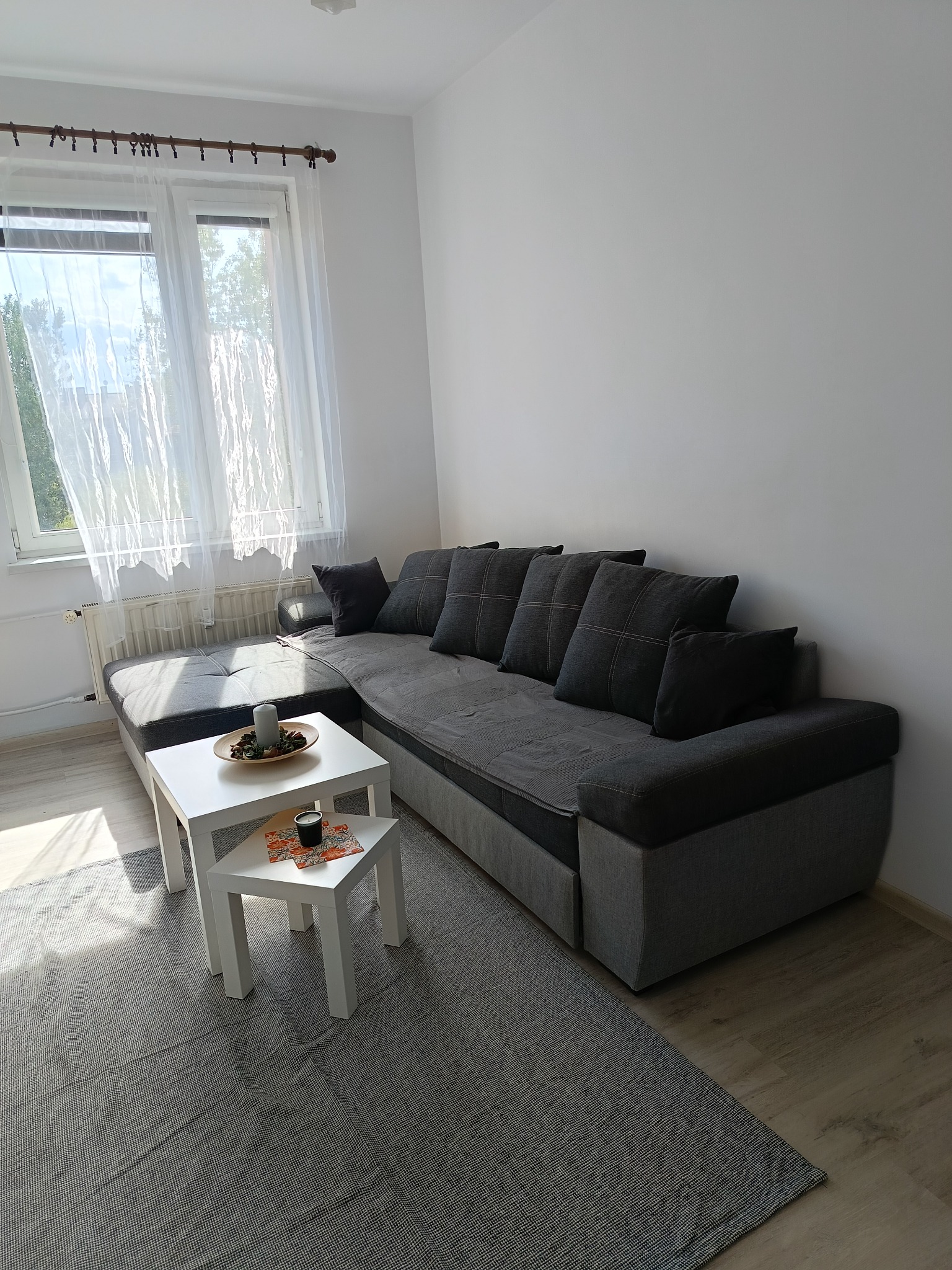 Jasne wnętrze pokoju z szarą, narożną kanapą, białym stolikiem i dywanem. Naturalne światło wpada przez okno z firankami. Widoczny fragment podłogi z paneli.