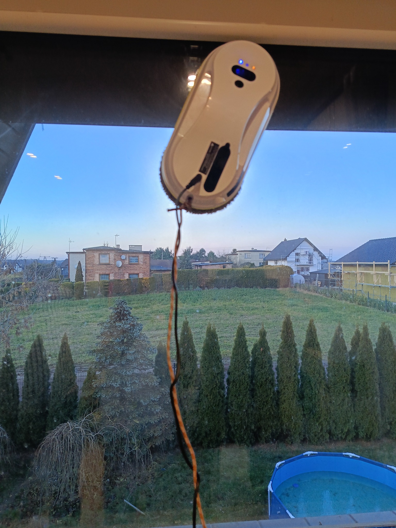 Robot do mycia okien w trakcie pracy na szybie z widokiem na ogród i domy. Urządzenie automatyczne, białe, z kablem zasilającym. Czyste okno.