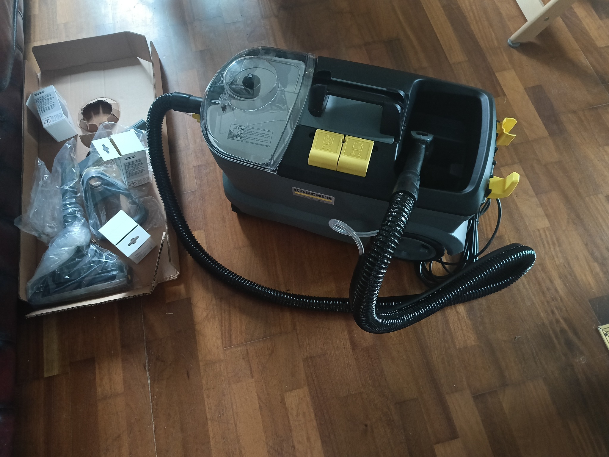 Rozpakowany odkurzacz piorący Karcher z akcesoriami w kartonie, gotowy do użycia na drewnianej podłodze. Widoczne detale urządzenia i elementy zestawu.