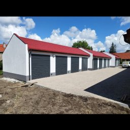 BARTOSZ BAŁDYGA BARTYNK - Rząd nowo wybudowanych garaży z czerwonymi dachami i szarymi bramami, świeżo otynkowane, z widocznym fragmentem terenu przed nimi.