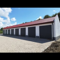 BARTOSZ BAŁDYGA BARTYNK - Rząd garaży z czerwonym dachem i szarymi bramami, widok z zewnątrz na kostkę brukową. Budynki otynkowane na biało, w słoneczny dzień.