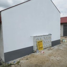 BARTOSZ BAŁDYGA BARTYNK - Świeżo pomalowana elewacja budynku z szarą podmurówką. Widoczne puszki z farbą i drabina. Biała ściana kontrastuje z ziemistym otoczeniem.