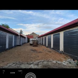 BARTOSZ BAŁDYGA BARTYNK - Rząd garaży z szarymi bramami i czerwonymi dachami, widok z perspektywy ulicy. Część garaży ma surowy tynk, inne są pomalowane. Na końcu uliczki stoi przyczepa kempingowa.