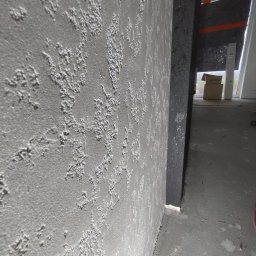 BARTOSZ BAŁDYGA BARTYNK - Szara ściana z dekoracyjną, ziarnistą strukturą tynku, widoczna z bliska pod kątem. W tle fragment okna i surowego stanu wykończenia wnętrza.