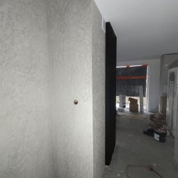 BARTOSZ BAŁDYGA BARTYNK - Ściana z dekoracyjną, szarą strukturą tynku w rogu pomieszczenia. Widoczny fragment instalacji elektrycznej i podłoga w trakcie prac wykończeniowych.