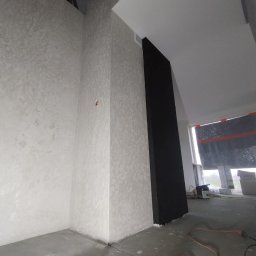 BARTOSZ BAŁDYGA BARTYNK - Wykończone wnętrze z dekoracyjną, jasną ścianą o fakturze i czarnym, pionowym elementem. Widoczny surowy beton na podłodze i okno z zasłoniętą szybą.