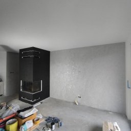 BARTOSZ BAŁDYGA BARTYNK - Pokój po gładziowaniu z czarnym kominkiem narożnym. Widoczne narzędzia i materiały malarskie. Ściana wykończona w odcieniach szarości z delikatnym połyskiem.