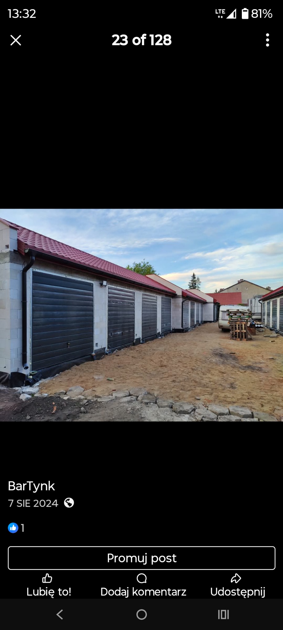 Rząd garaży z bramami rolowanymi w kolorze antracytu, wykończonych czerwoną dachówką. Budynki w stanie surowym, z białych bloczków. Widok z zewnątrz, w słoneczny dzień.