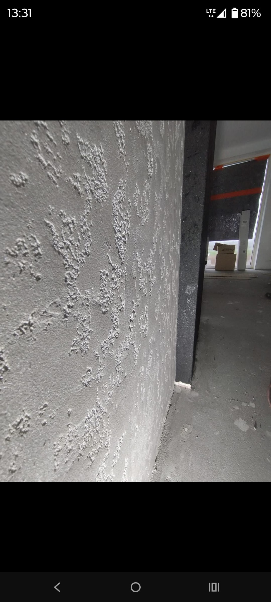 Szara ściana z dekoracyjną, ziarnistą strukturą tynku, widoczna z bliska pod kątem. W tle fragment okna i surowego stanu wykończenia wnętrza.