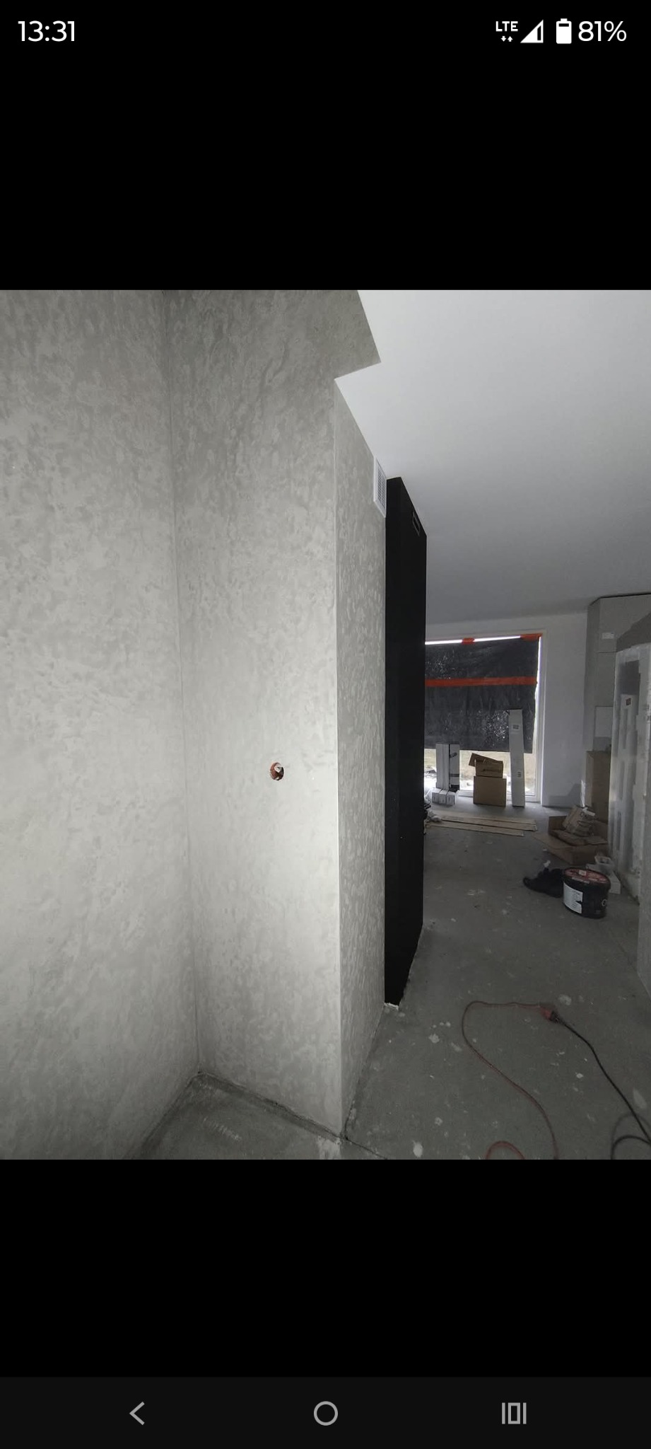 Ściana z dekoracyjną, szarą strukturą tynku w rogu pomieszczenia. Widoczny fragment instalacji elektrycznej i podłoga w trakcie prac wykończeniowych.