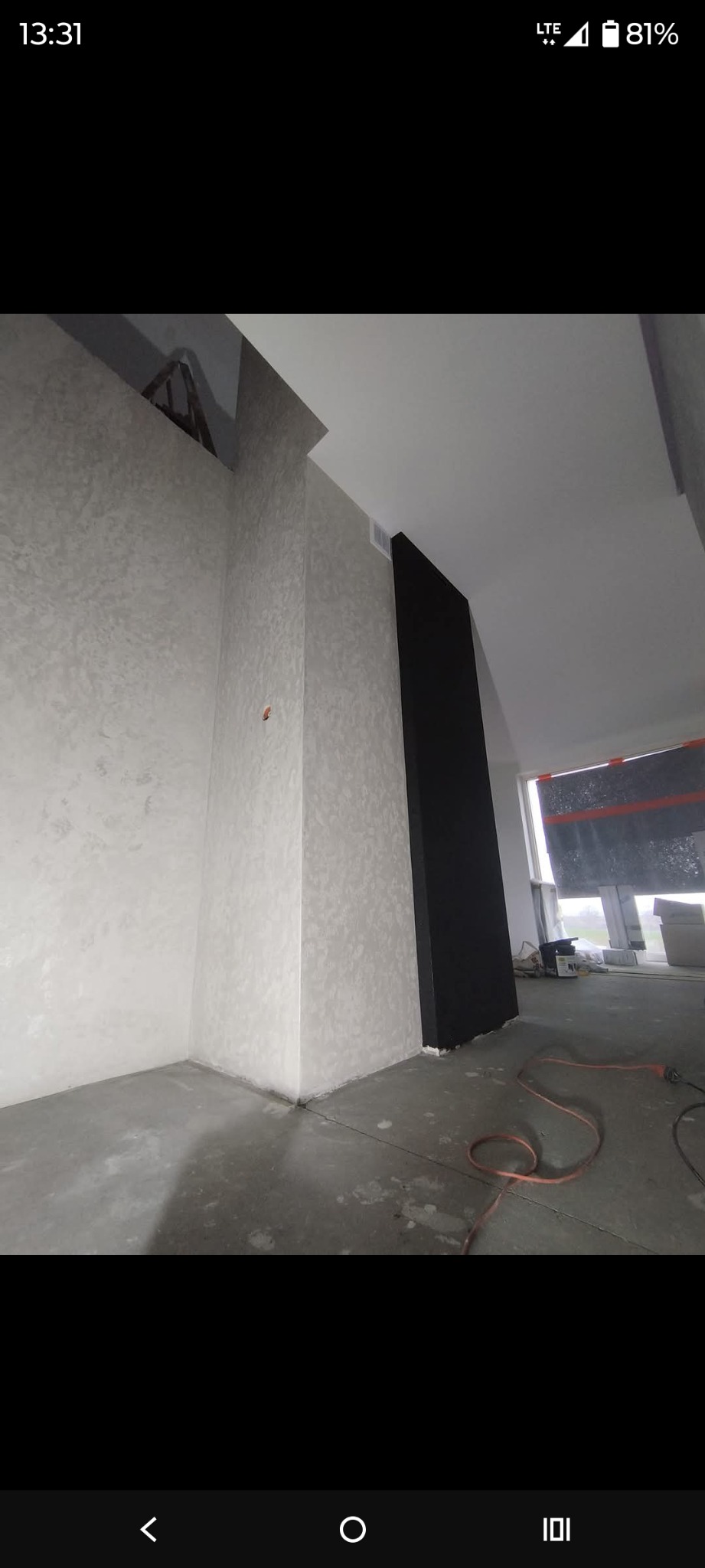 Wykończone wnętrze z dekoracyjną, jasną ścianą o fakturze i czarnym, pionowym elementem. Widoczny surowy beton na podłodze i okno z zasłoniętą szybą.