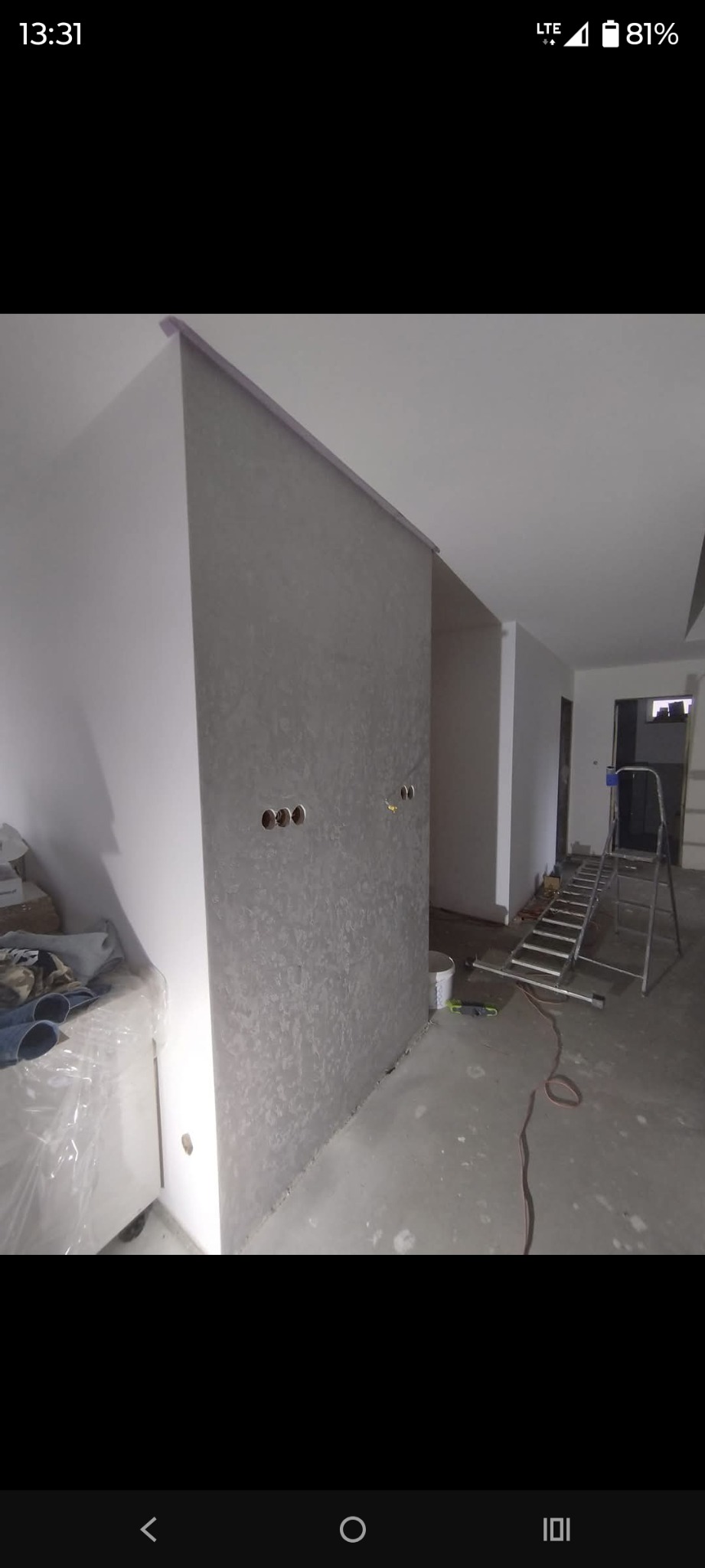 Ściana z szarą, dekoracyjną strukturą, widoczne otwory na gniazdka elektryczne. W tle drabina i przygotowane pomieszczenie do dalszych prac wykończeniowych.