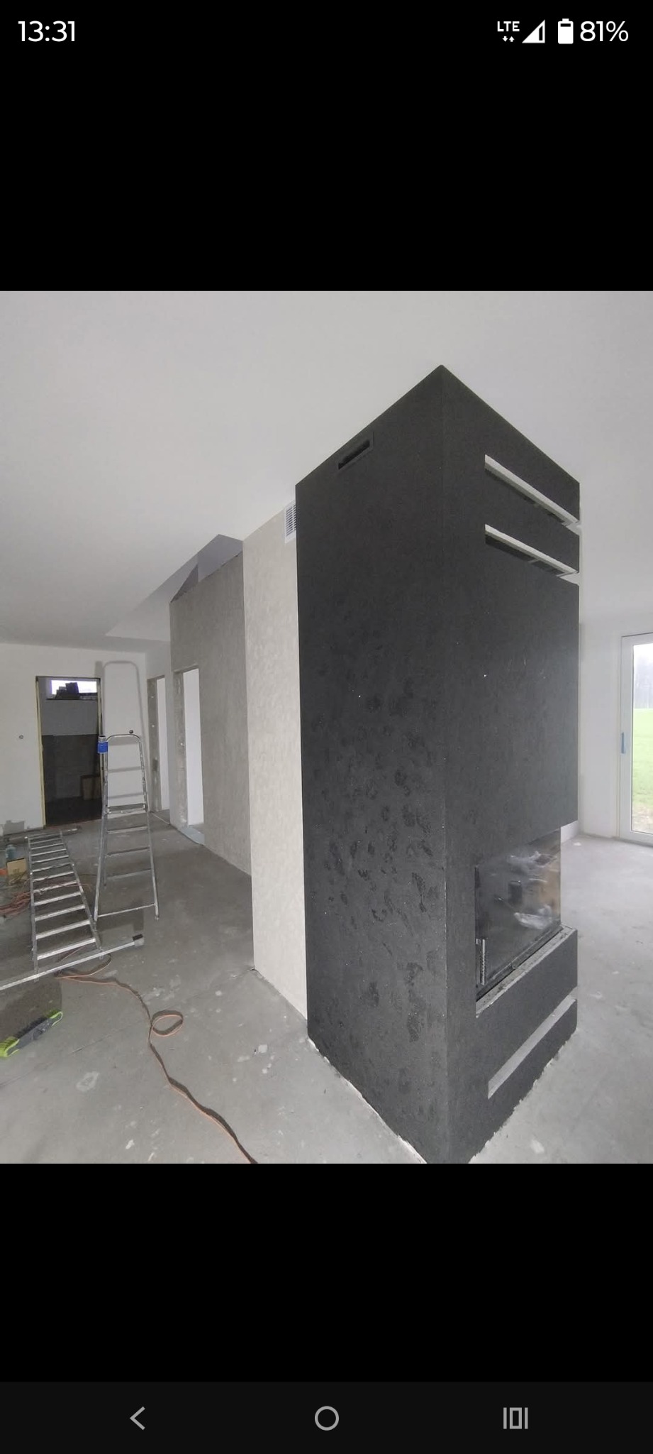 Wnętrze w trakcie wykańczania: czarny kominek z poziomymi wnękami, ściany z fakturą, drabina, surowa podłoga. Widok na okno i drzwi.