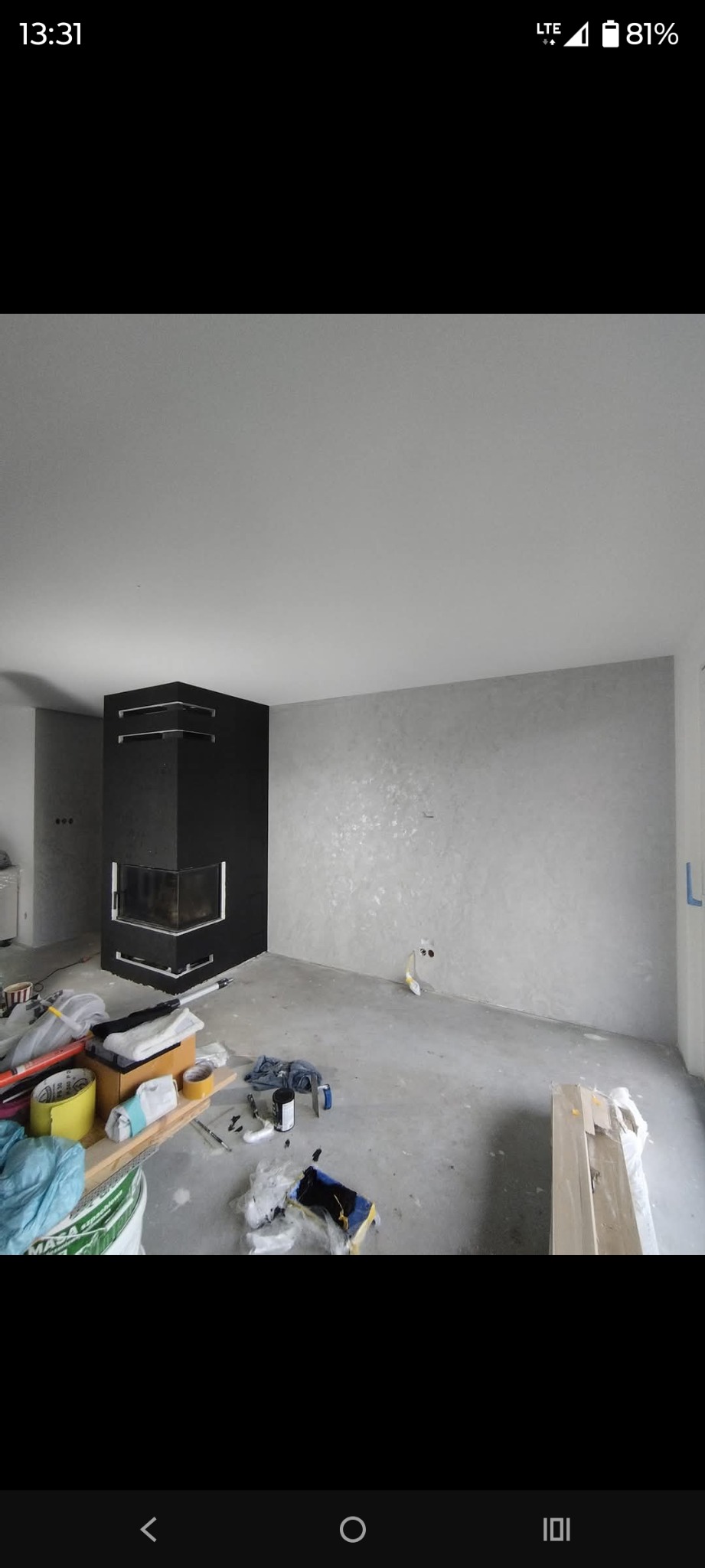 Pokój po gładziowaniu z czarnym kominkiem narożnym. Widoczne narzędzia i materiały malarskie. Ściana wykończona w odcieniach szarości z delikatnym połyskiem.