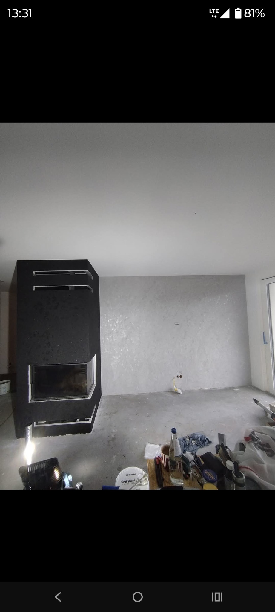 Wnętrze w trakcie wykańczania: czarny kominek o geometrycznym kształcie, ściana z dekoracyjną fakturą, narzędzia i materiały budowlane na podłodze. Surowy, nowoczesny styl.