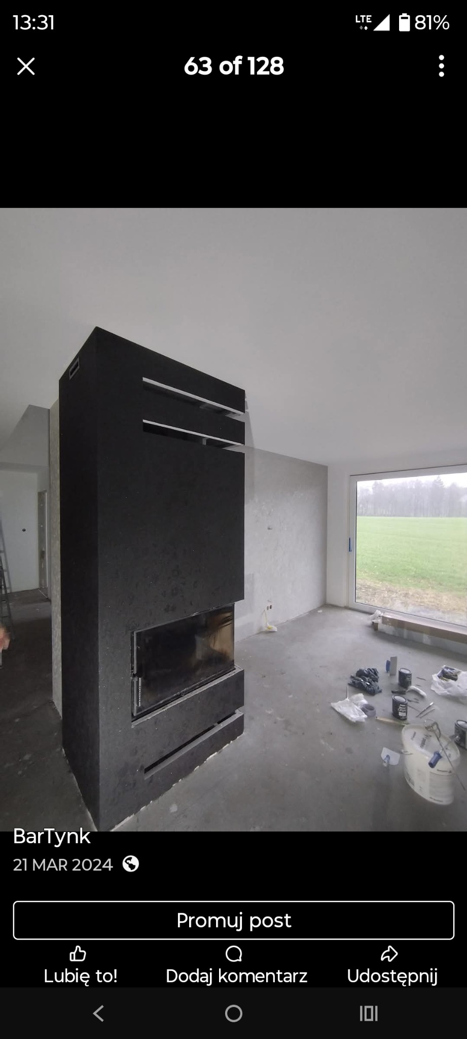 Nowoczesny, czarny kominek z przeszklonym paleniskiem w surowym wnętrzu. Widoczne narzędzia i materiały budowlane, co sugeruje etap wykończeniowy. Duże okno z widokiem na zieleń.