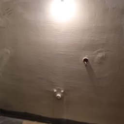 Instal & Serwis Jakub Sikora - Ściana w trakcie remontu łazienki z widocznymi otworami na instalacje i częściowo położoną gładzią. Widoczne elementy instalacji wodnej i elektrycznej.