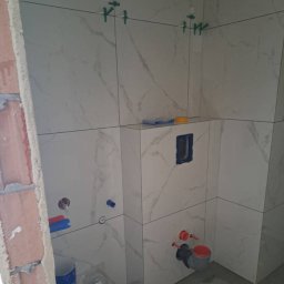 Instal & Serwis Jakub Sikora - Świeżo ułożone, marmurowe płytki w łazience w Krakowie, z widocznymi elementami instalacji wodnej i poziomującymi klipsami. W tle surowa ściana z cegły.