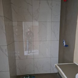 Instal & Serwis Jakub Sikora - Wnętrze łazienki w trakcie prac. Widoczne płytki imitujące marmur na ścianach, przygotowane miejsce na brodzik i fragmenty płytek na podłodze. Surowy stan wykończenia.