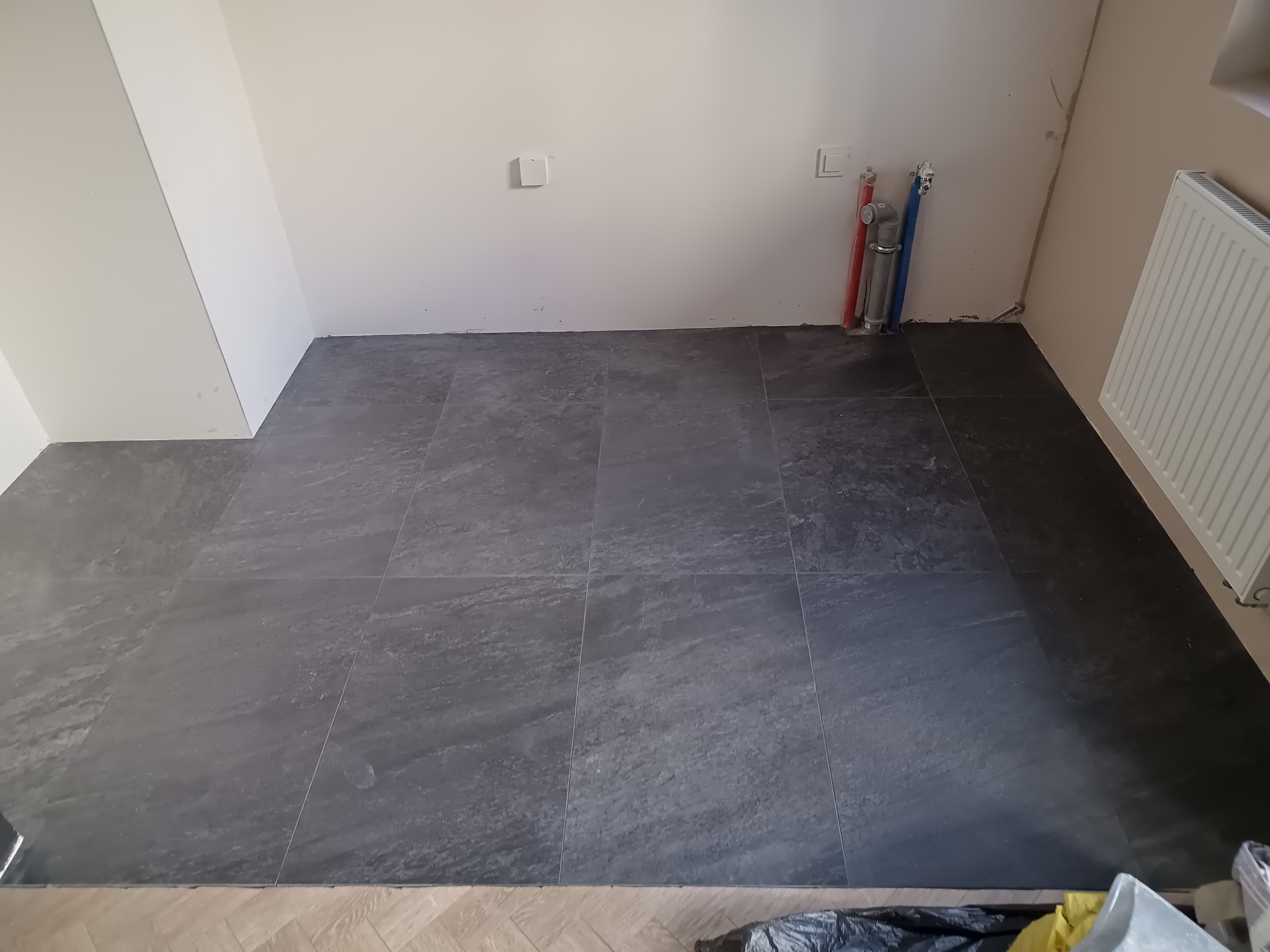 Ułożona, ciemnoszara podłoga z płytek w pomieszczeniu z białymi ścianami i grzejnikiem. Widoczne rury instalacyjne przy ścianie. Fragment podłogi z parkietu w dolnym rogu.