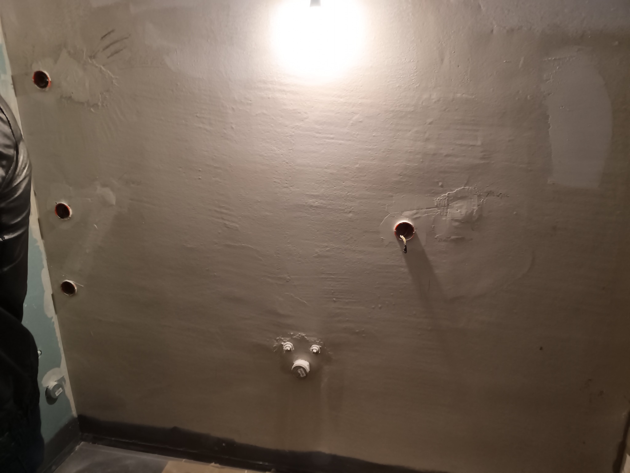Ściana w trakcie remontu łazienki z widocznymi otworami na instalacje i częściowo położoną gładzią. Widoczne elementy instalacji wodnej i elektrycznej.