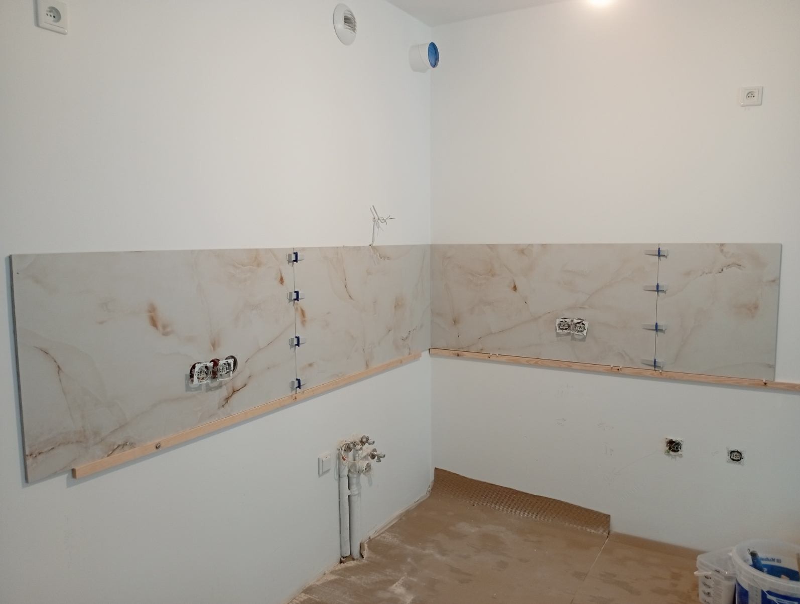 Fragment ściany z zamontowanymi płytkami imitującymi marmur, zabezpieczonymi niebieskimi klipsami, wspartymi na drewnianej listwie. Widoczne gniazdka elektryczne i rury.