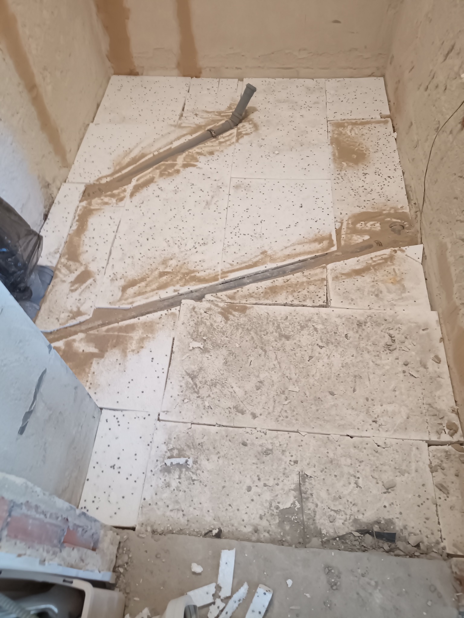 Fragment łazienki w trakcie prac. Widoczne białe płyty podłogowe z drobnymi otworami, częściowo zabrudzone zaprawą, oraz szara rura odpływowa. Ściany surowe, w trakcie przygotowania.