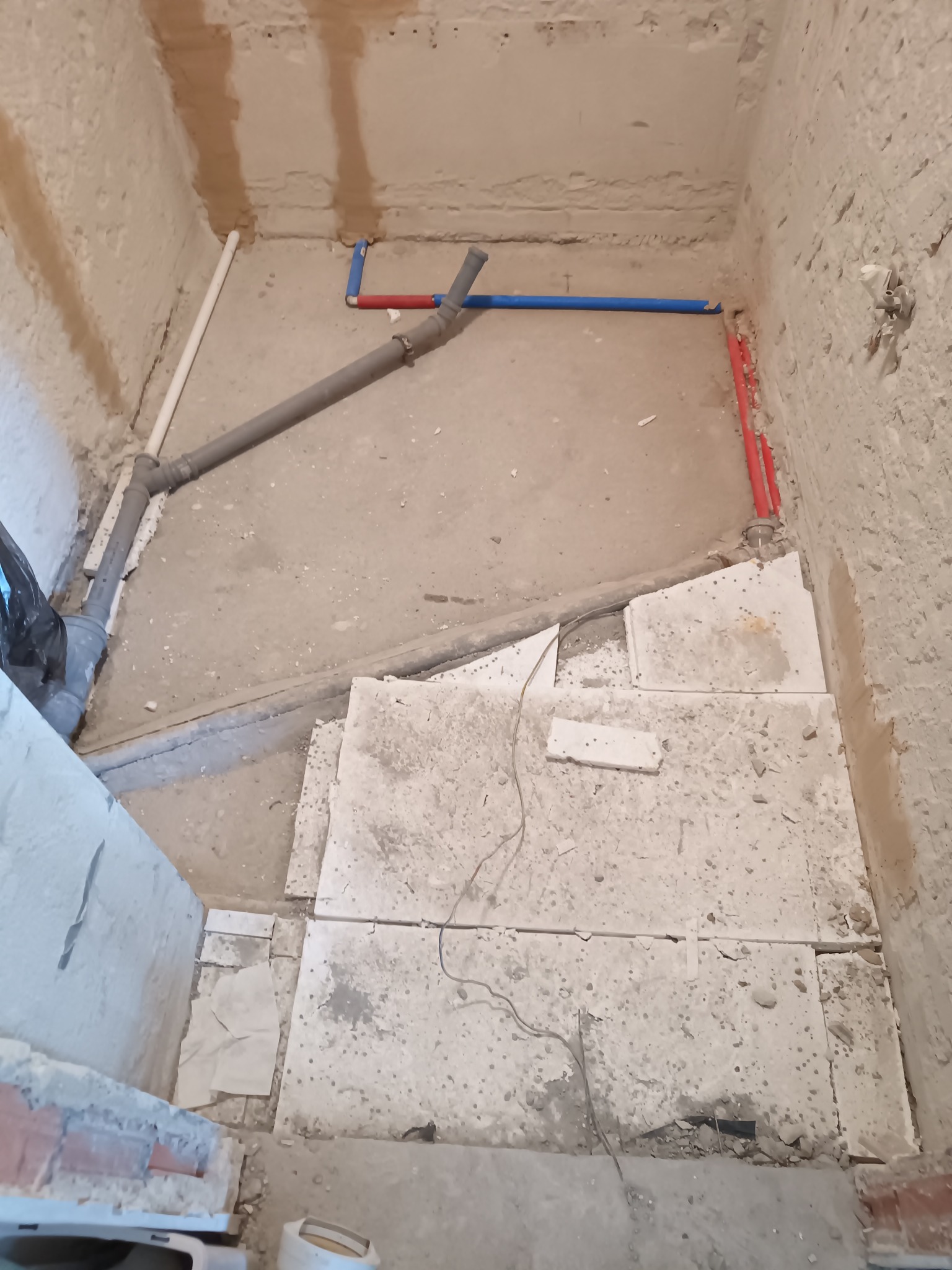 Surowy stan łazienki w trakcie prac: widoczne rury wodne (czerwone, niebieskie), kanalizacyjne (szare) oraz przygotowane podłoże pod posadzkę. Ściany w stanie deweloperskim.