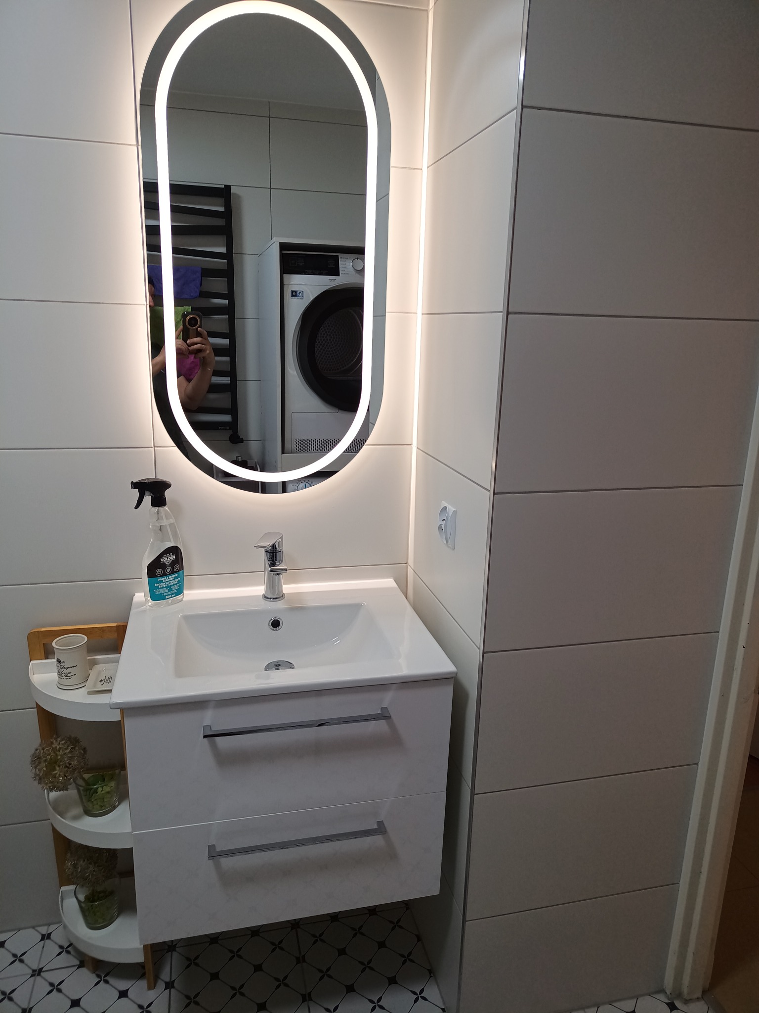 Biała umywalka z szafką i podświetlanym lustrem w owalnej ramie. W tle pralka i grzejnik drabinkowy. Minimalistyczna łazienka z geometryczną podłogą.