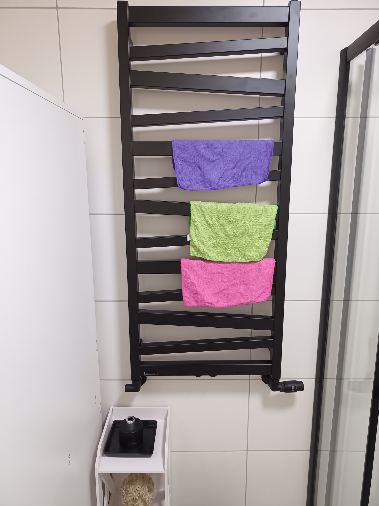 Nowoczesna łazienka z czarnym grzejnikiem drabinkowym, na którym wiszą trzy kolorowe ręczniki: fioletowy, zielony i różowy. Białe płytki na ścianach i minimalistyczna szafka.