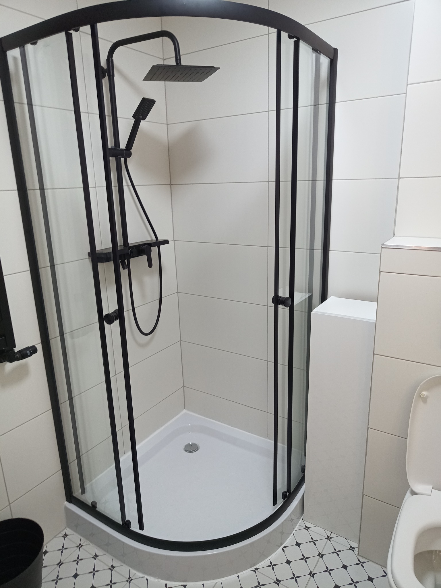 Nowoczesna kabina prysznicowa z czarnymi profilami i deszczownicą, białe płytki na ścianach, wzorzysta podłoga. Minimalistyczny design łazienki.