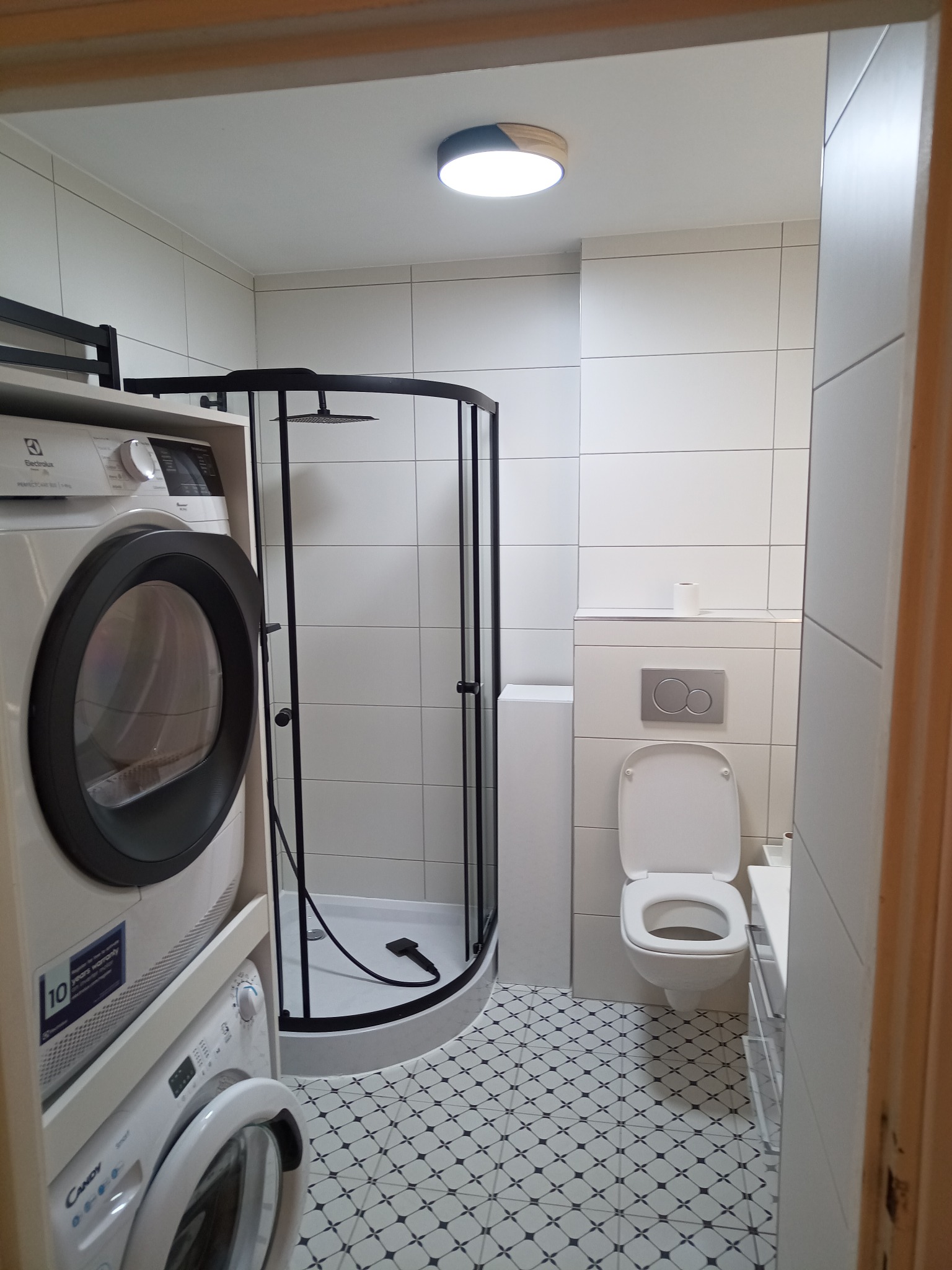 Nowoczesna łazienka po remoncie: białe płytki, kabina prysznicowa z czarnymi ramami, toaleta i pralka z suszarką w zabudowie. Podłoga w biało-czarne wzory.