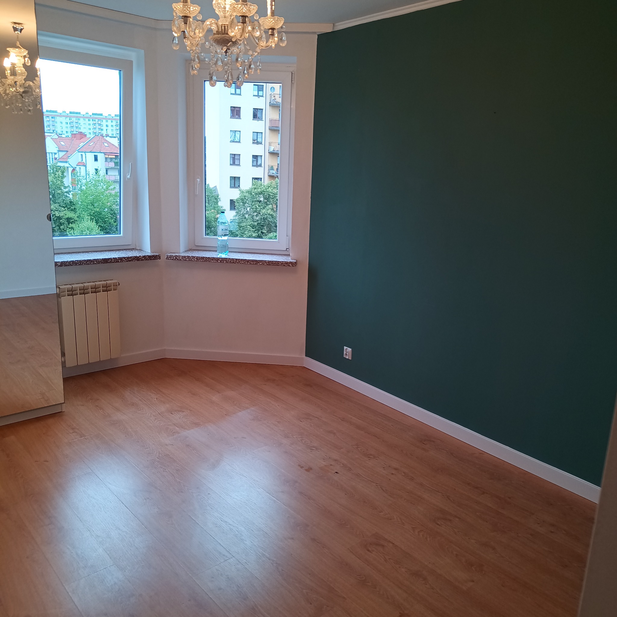 Pomalowane na ciemnozielono ściany w pokoju z parkietem, widok na zewnątrz przez okna z białymi ramami. Żyrandol kryształowy w górnej części kadru.