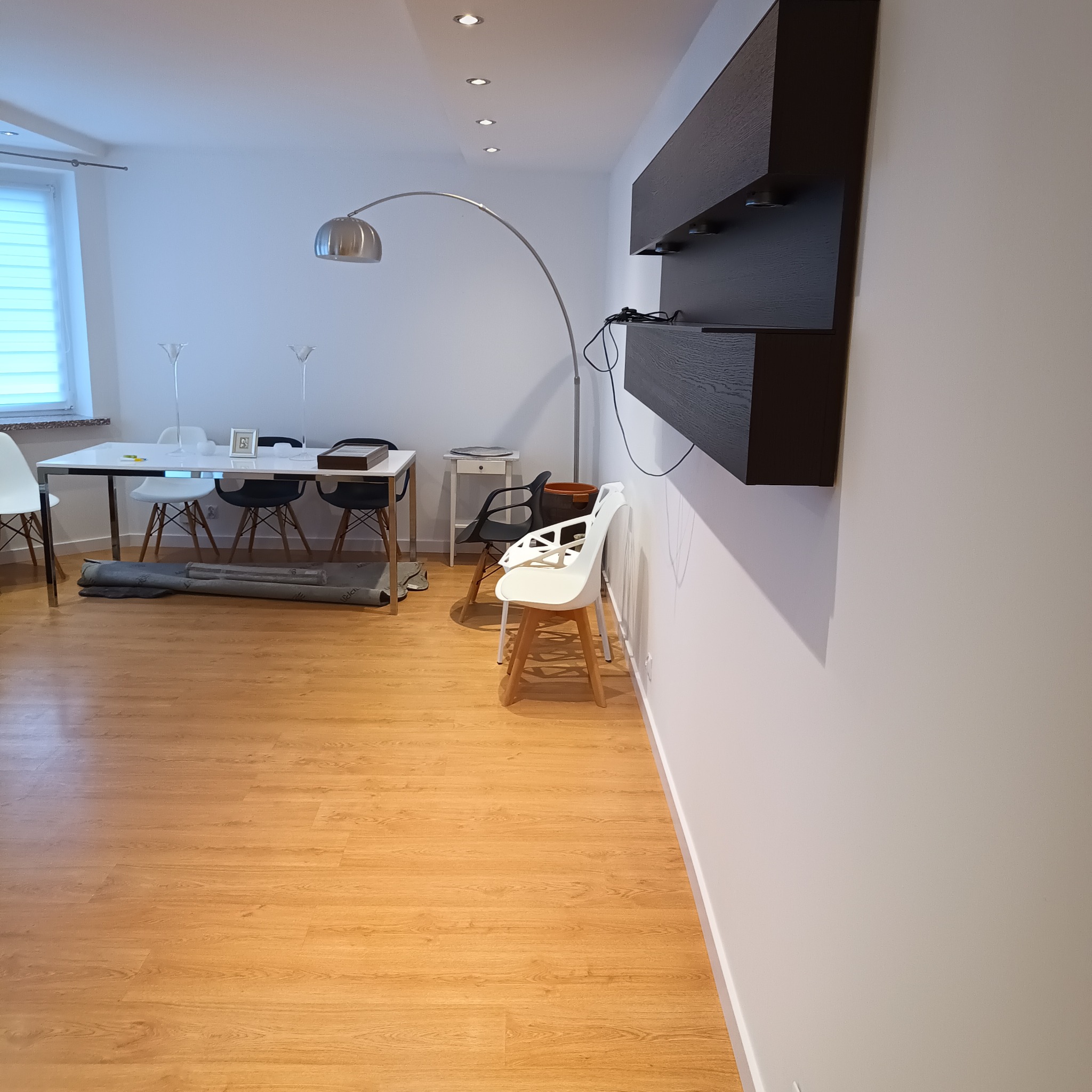 Jasne wnętrze z drewnianą podłogą, białymi ścianami i nowoczesnymi meblami. Na ścianie wisi ciemna, drewniana półka. Widoczny stół z krzesłami i lampa łukowa.