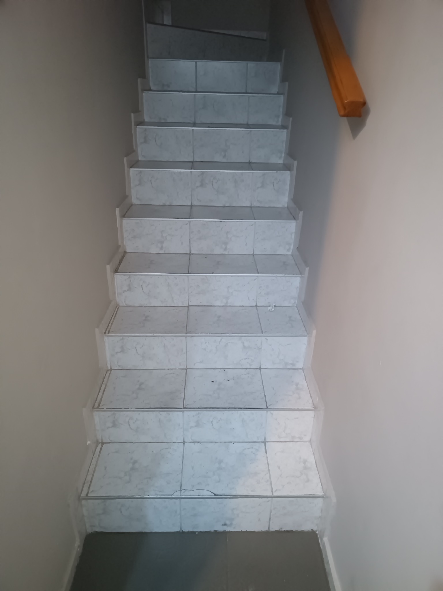 Wykończone schody z jasnymi, marmurkowymi płytkami, widziane z góry. Drewniana poręcz po prawej stronie. Ściany w neutralnym odcieniu beżu.