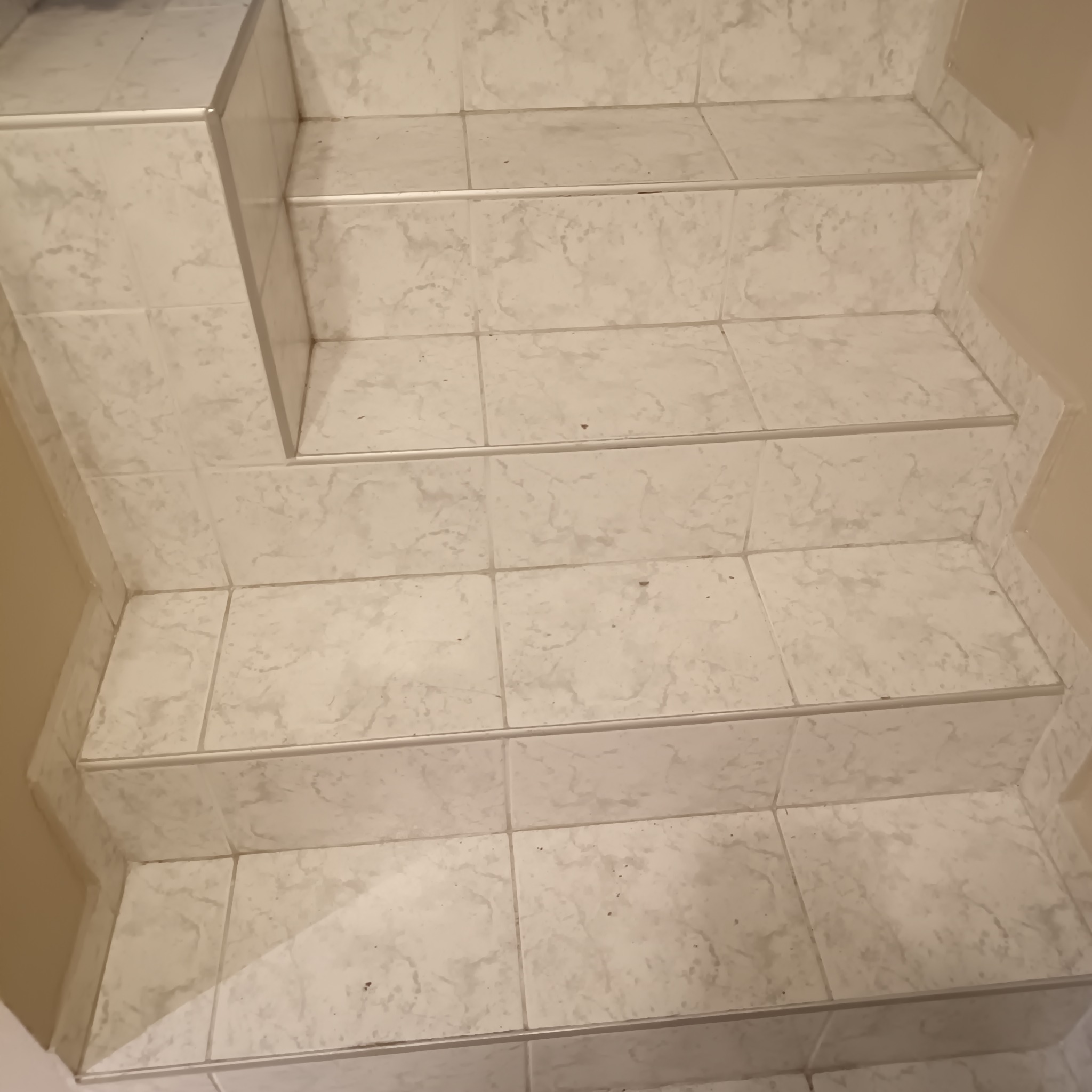 Wykończone schody z jasnych, marmurkowych płytek, zabezpieczone srebrnymi listwami. Widok z góry, ukazujący precyzję ułożenia i estetyczne detale wykończenia.