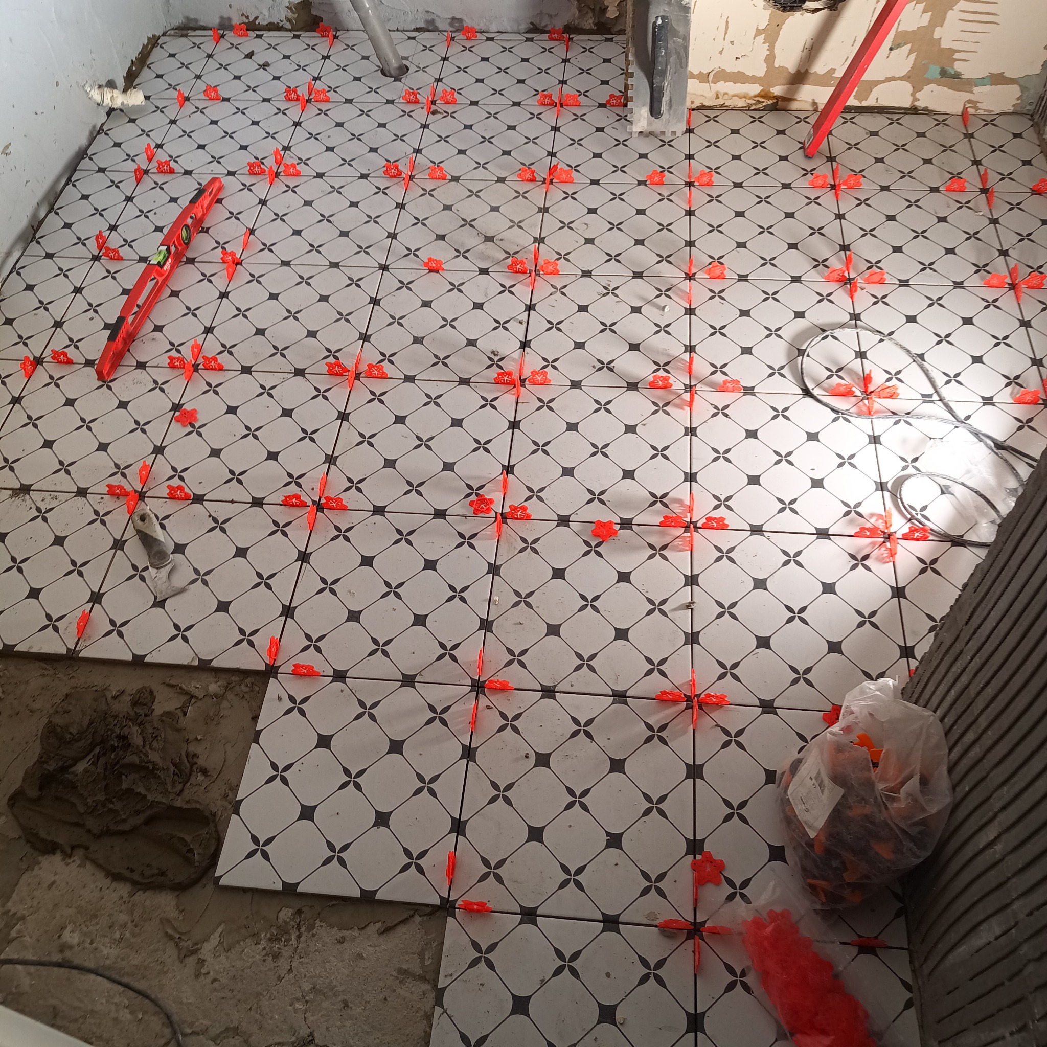 Układanie podłogi z płytek w romby, z użyciem czerwonych krzyżyków dystansowych i poziomicy. Częściowo położone płytki, widoczny klej i zapas krzyżyków.