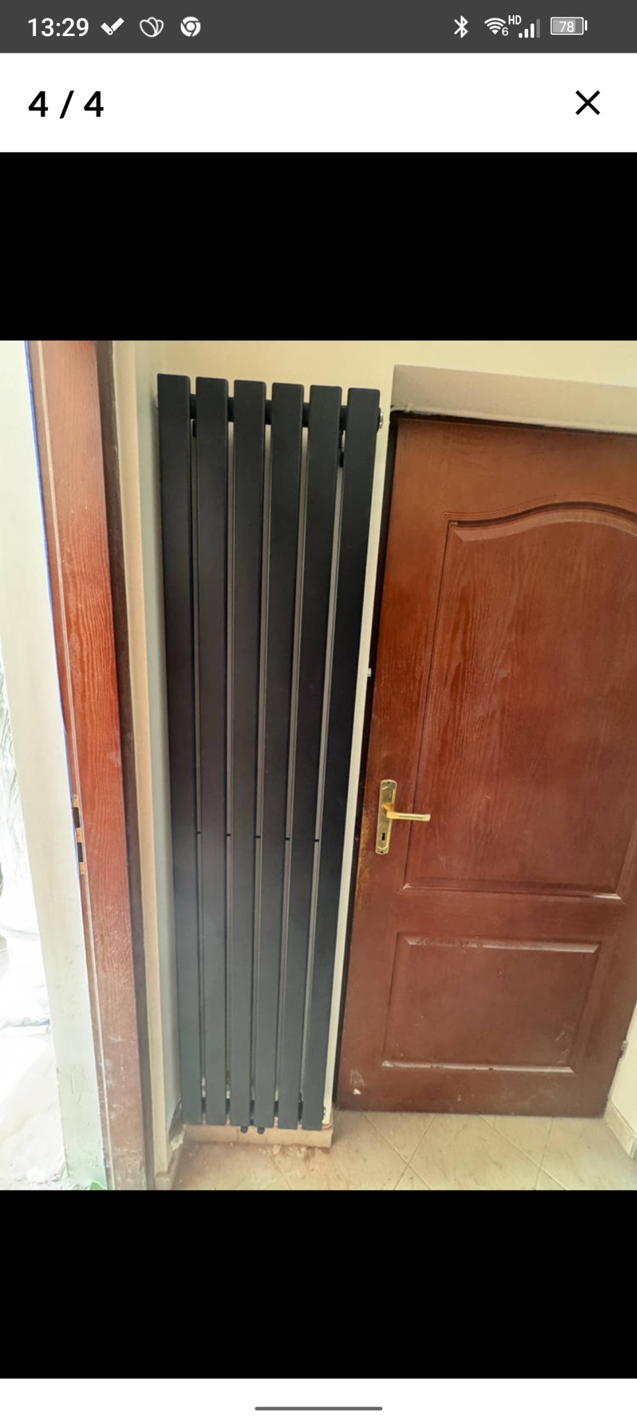 Zamontowany, pionowy, czarny grzejnik panelowy w wąskim korytarzu, obok brązowych drzwi. Widoczne fragmenty ościeżnic i podłogi z płytek.