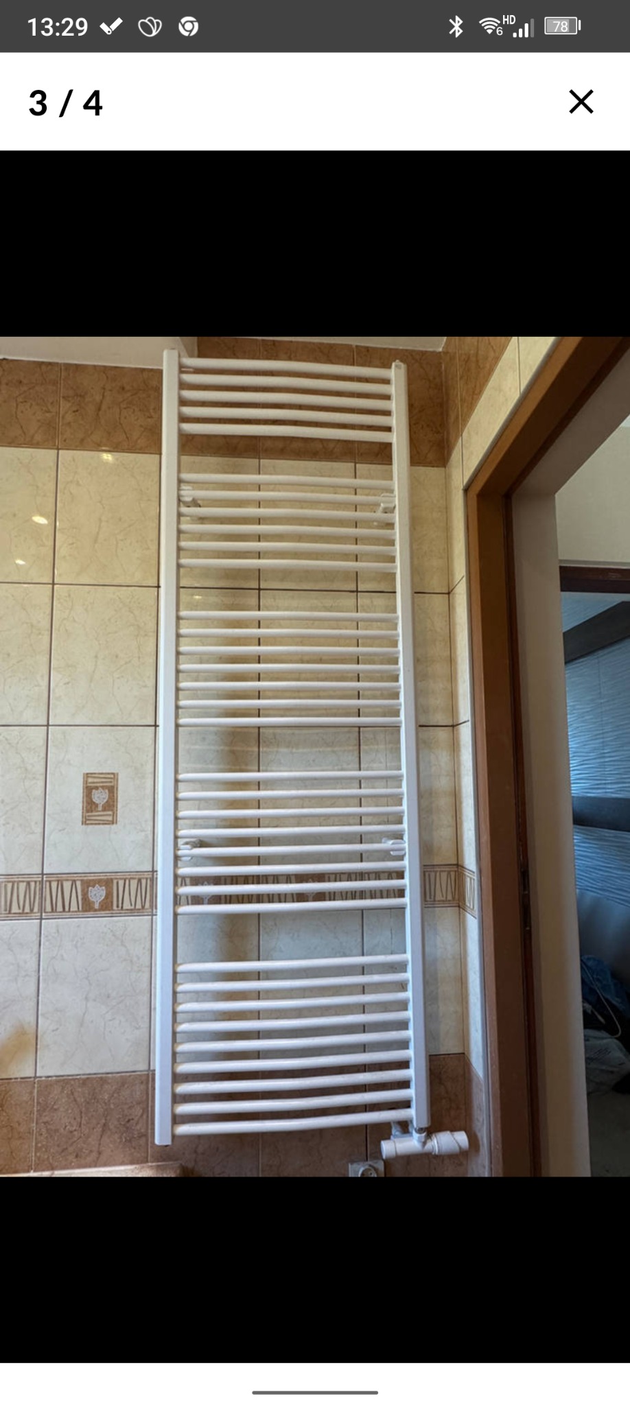 Biały, drabinkowy grzejnik łazienkowy zamontowany na ścianie z beżowymi płytkami. Widoczny fragment drzwi. Ujęcie z bliska, prezentujące detale instalacji.