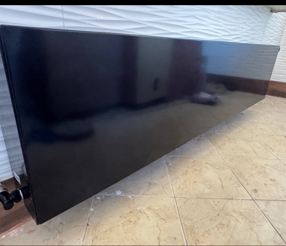 Czarny, błyszczący grzejnik dekoracyjny na tle białej, teksturowanej ściany i beżowej podłogi z płytek. Widoczne przyłącza grzejnika.