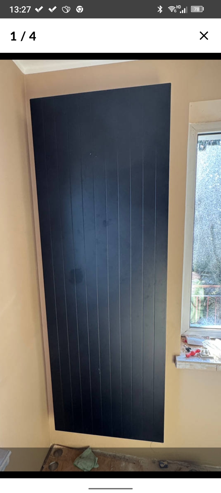 Ciemna, pionowa płyta z wyraźnymi żłobieniami na beżowej ścianie, obok okna z widokiem na zewnątrz. W rogu podłogi widoczny fragment podkładu i szmatka.