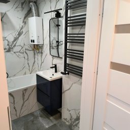 Sergi_job - Wykończona łazienka z marmurowymi płytkami, zabudowaną umywalką w kolorze granatowym i czarnym grzejnikiem drabinkowym. Widoczny bojler i białe drzwi.